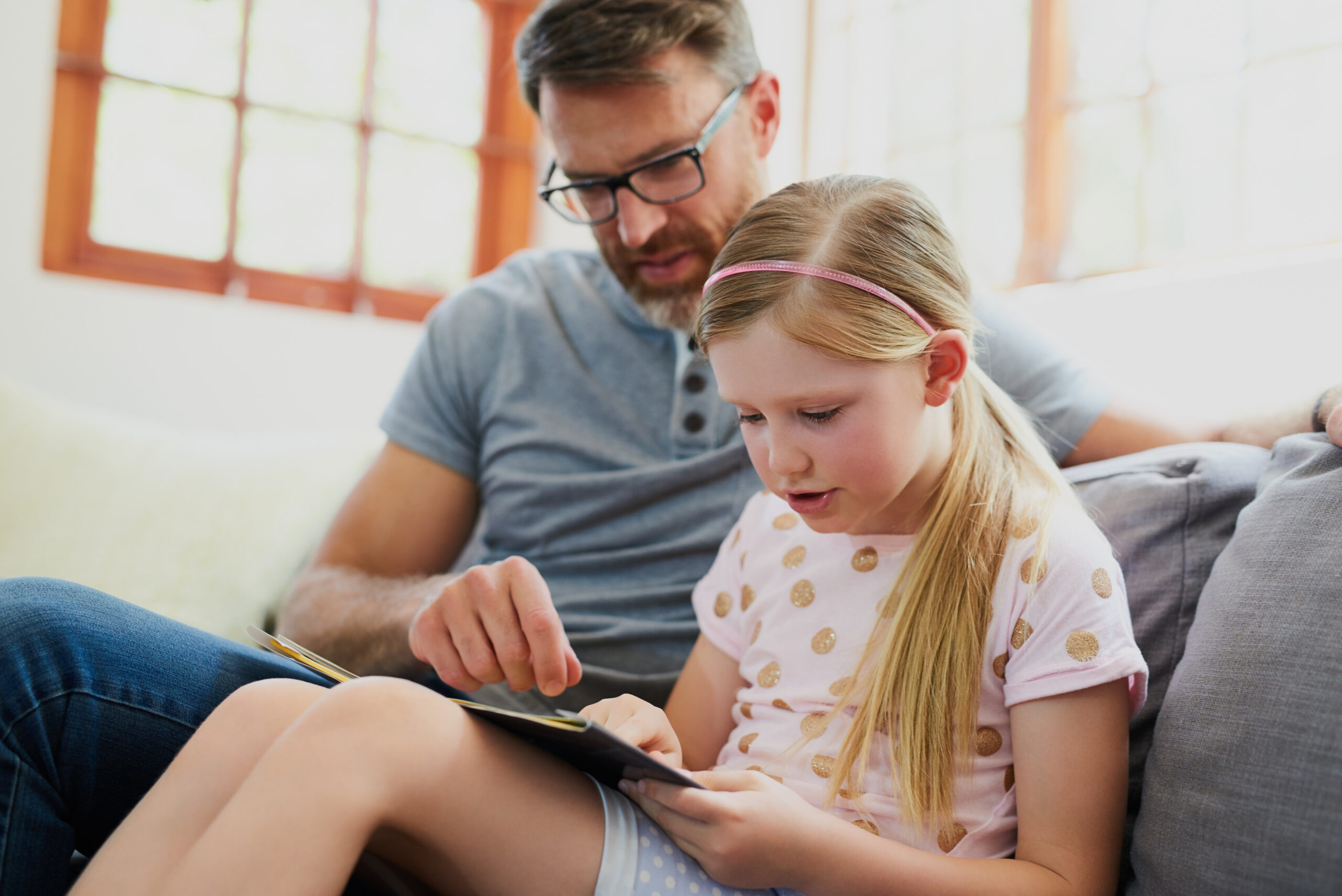 Father, reading and daughter with book on sofa for story, learning or education at home. Dad, child or kid with novel for literature, language or helping with pronunciation on lounge couch at house